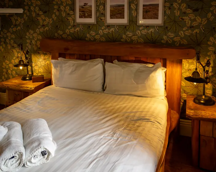 A double bed with a rough-hewn oak headboard, brass reading lamps and oak-leaf wallpaper beneath framed moorland prints